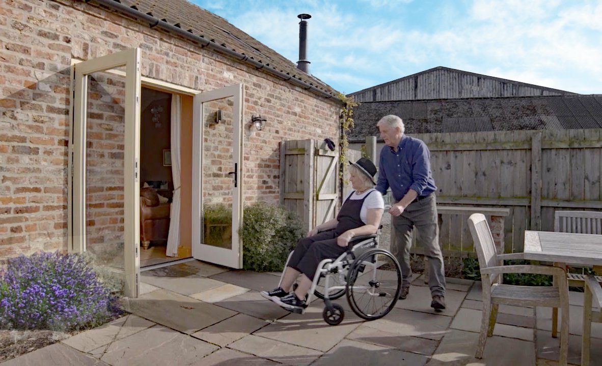 Man assists woman in wheelchair outside house.